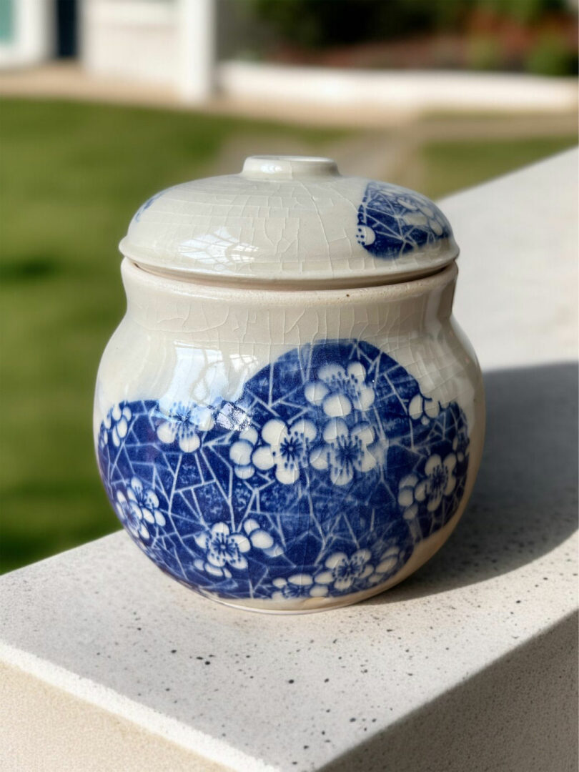 A ceramic jar with a lid, decorated with blue and white floral patterns in the style of Mark Jupiter, sits on a light-colored outdoor ledge with a grassy background.