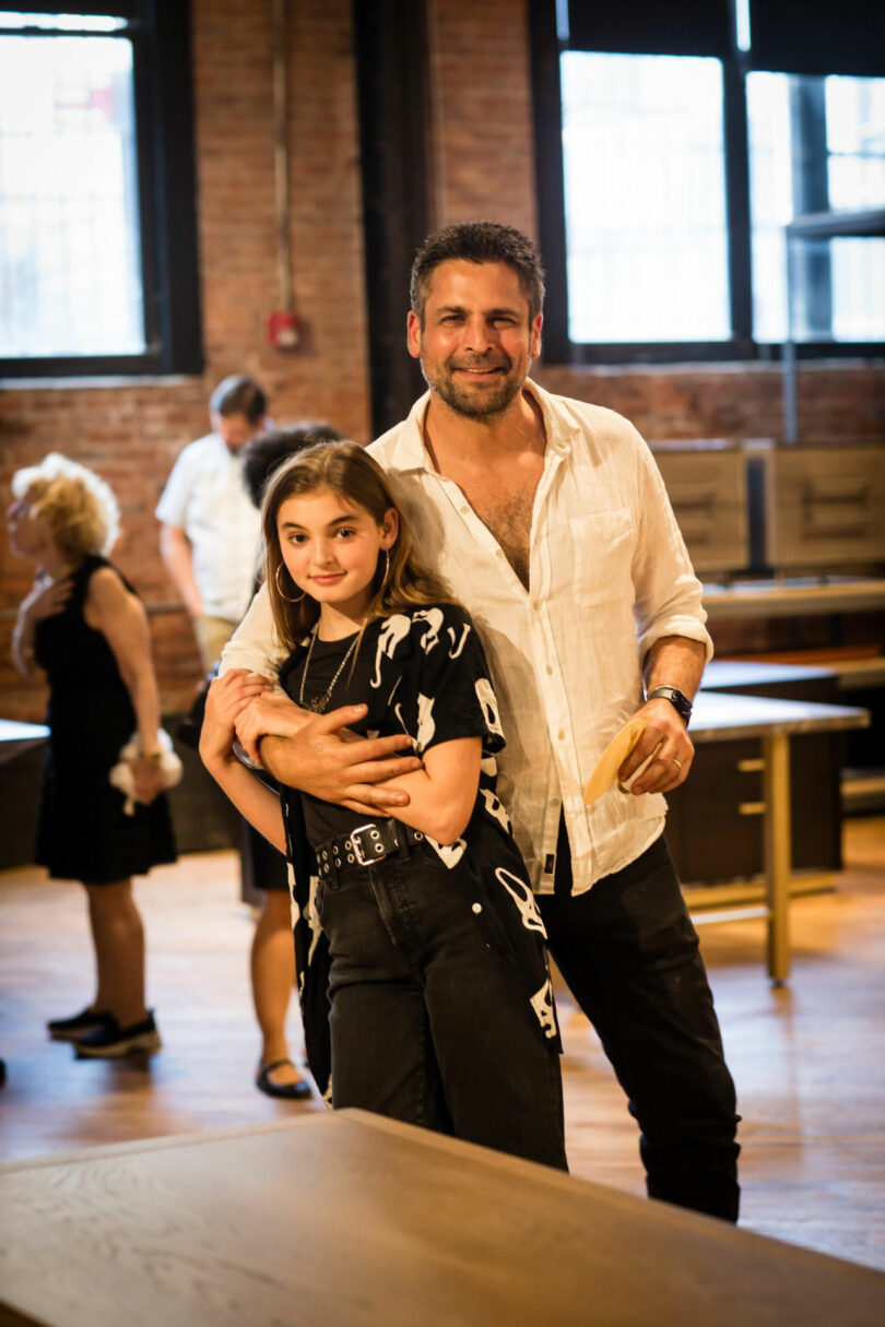 A man stands behind a young girl with his arms around her in a sunlit room with wooden floors and tables at Mark Jupiter. Other people are visible in the background.