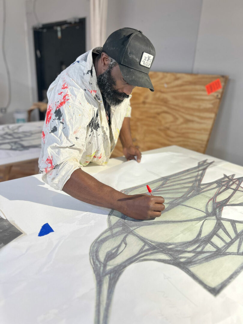 A man in a paint-splattered hoodie and cap, reminiscent of Mark Jupiter’s creative style, sketches abstract geometric shapes on a large sheet of paper in a studio setting.