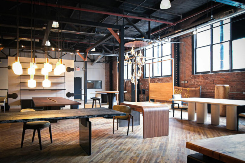 A modern industrial showroom by Mark Jupiter features various wooden tables and chairs, exposed brick walls, large windows, and hanging light fixtures.