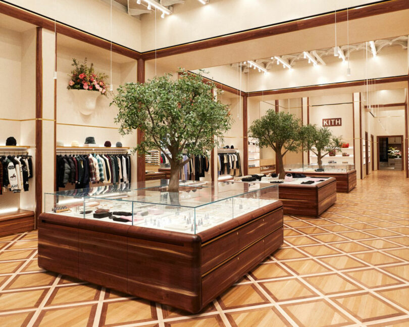 Modern boutique clothing store interior featuring Mark Jupiter wooden display cases with glass tops, indoor trees, and neatly arranged apparel on racks and shelves.