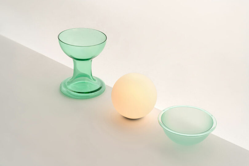 A green glass goblet, a glowing sphere reminiscent of the Gelato Lamp by Carlo Nason, and a shallow green glass bowl are arranged on a minimalist white and gray surface.