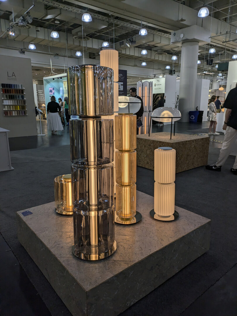 A display of cylindrical lamps with glass and metal finishes on a platform at an indoor exhibition, surrounded by booths and visitors.