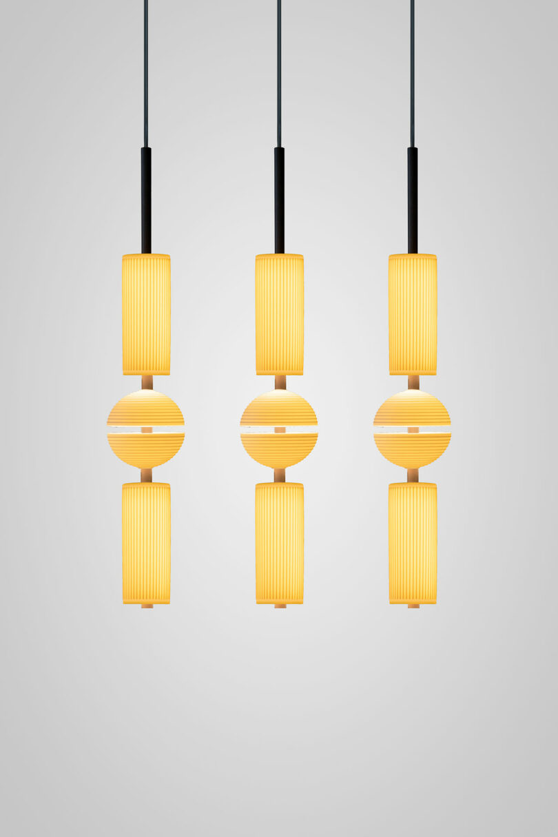 Three modern Lee Broom pendant lights with cylindrical and spherical yellow lampshades, suspended from black cords against a plain light gray background.