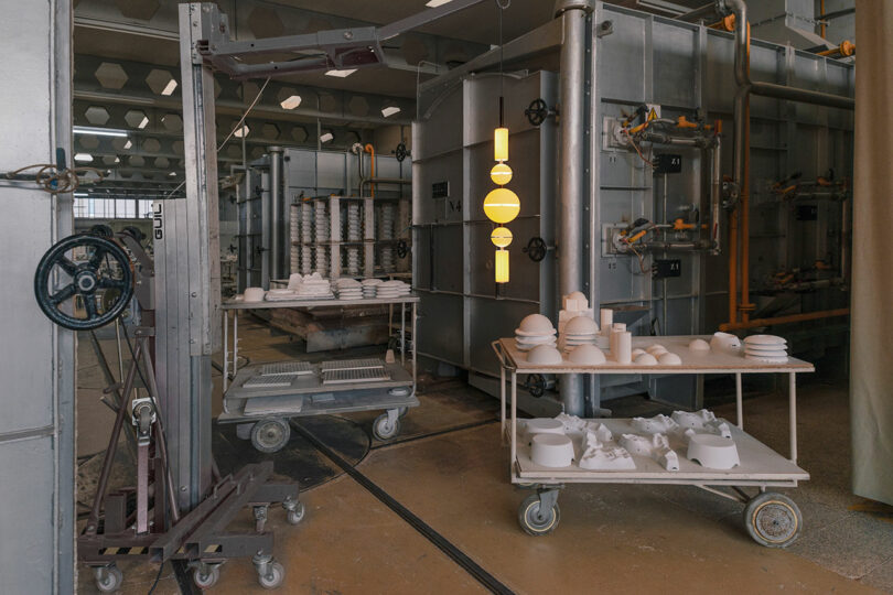 Industrial ceramics workshop with metal carts holding white ceramic molds and objects, surrounded by machinery and pipes—echoing the refined yet industrial aesthetic of Lee Broom’s innovative designs.