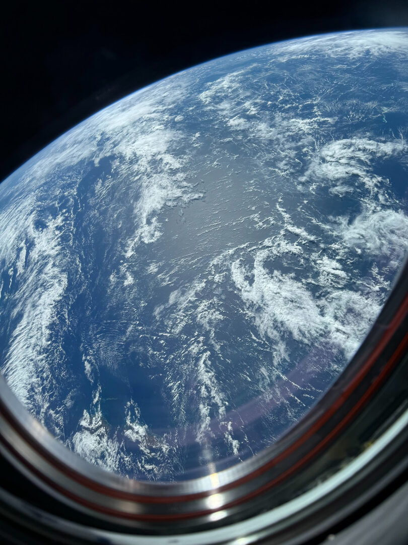 The photograph shows Earth from space, with a curved horizon and scattered clouds, seen through a round spacecraft window.