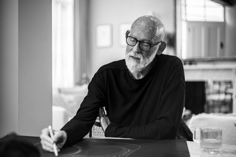 Elderly man with a beard and glasses, seated at a table, writes on a digital tablet with a stylus in a softly lit room.