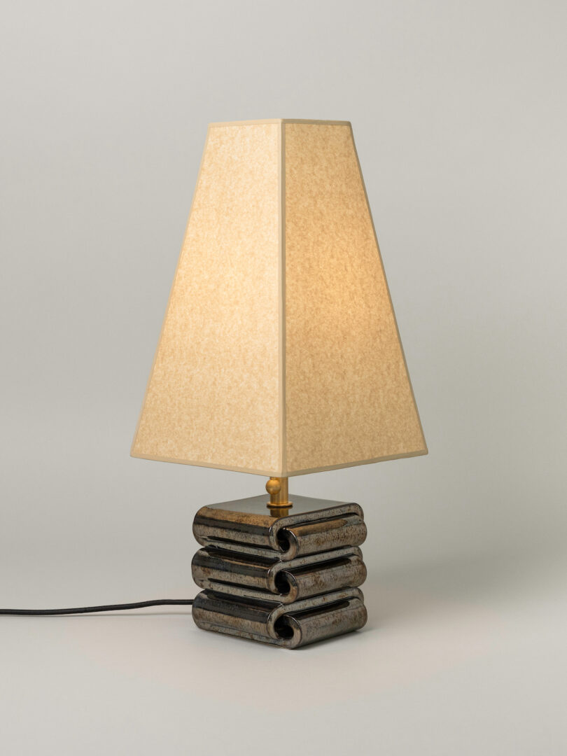 A table lamp with a beige, angular lampshade and a stacked, rectangular ceramic base with a dark, glossy finish. The lamp is plugged in and turned on, adding a touch of Virginia Sin lighting style to any room