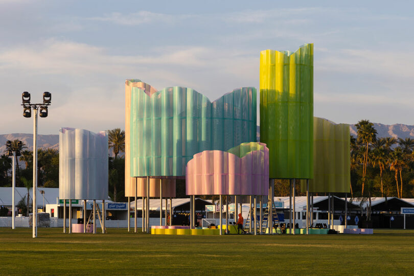 Large, colorful, translucent structures in pastel shades stand on a grassy field with tents and palm trees in the background—inviting you to Take Five and unwind amidst this vibrant scene.