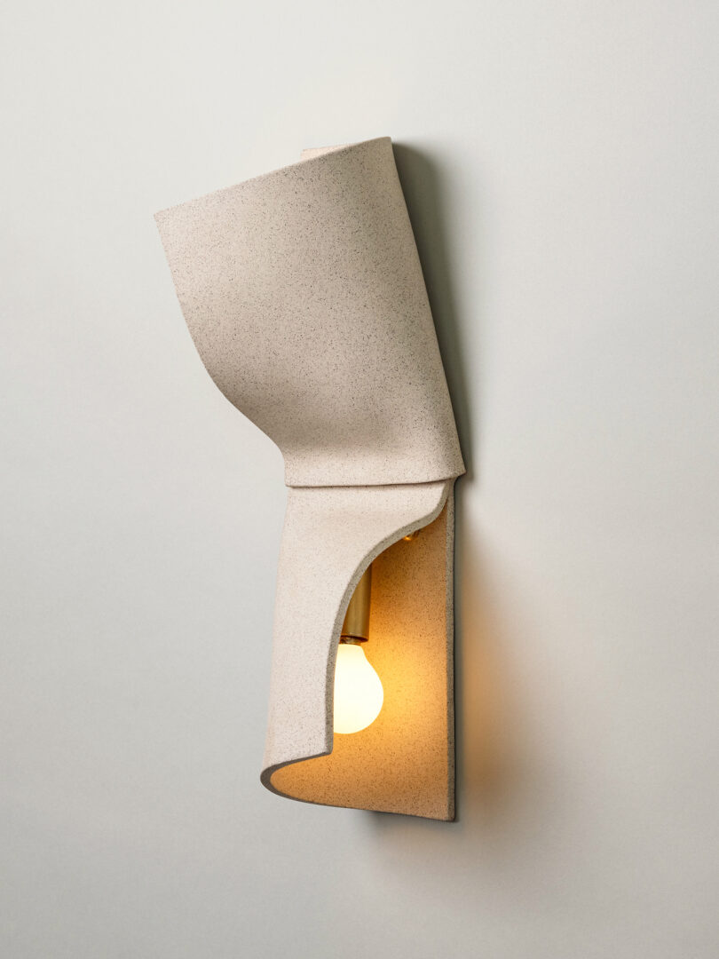 A wall-mounted Virignia Sin lighting fixture featuring a beige, folded, textured cover that partially reveals a round light bulb on a brass base