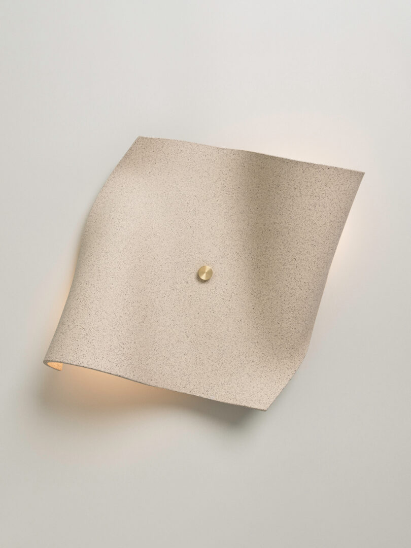 A beige, textured wall sconce with a slightly curved, square-shaped design and a small round central fixture evokes the artistry of Virginia Sin lighting, mounted elegantly on a white wall