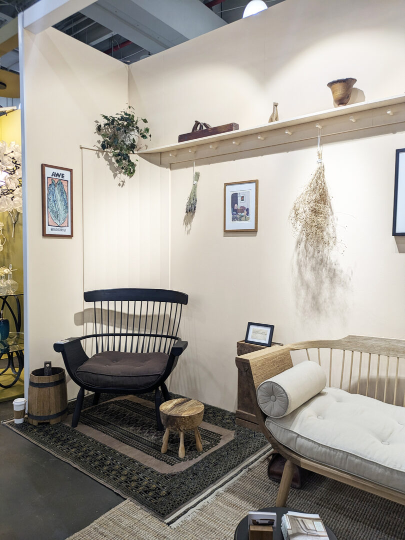 A cozy living space with a black wooden chair, small stool, beige daybed, wall shelf with decor, framed art, and hanging dried plants on light-colored walls.