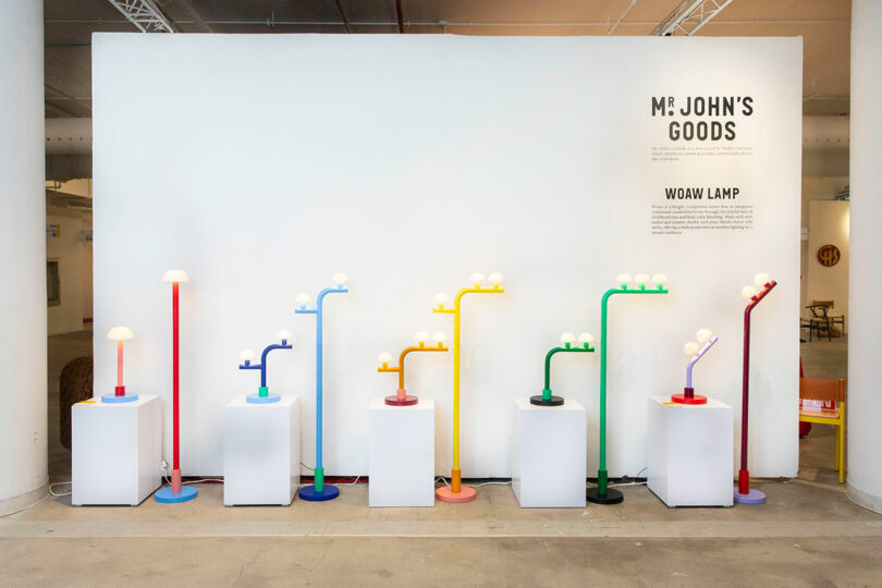 A display of colorful lamps in blue, yellow, green, and red—curated by Nader Gammas—are arranged on white pedestals in front of a wall with text reading "MR. JOHN'S GOODS" and "WOAW LAMP.