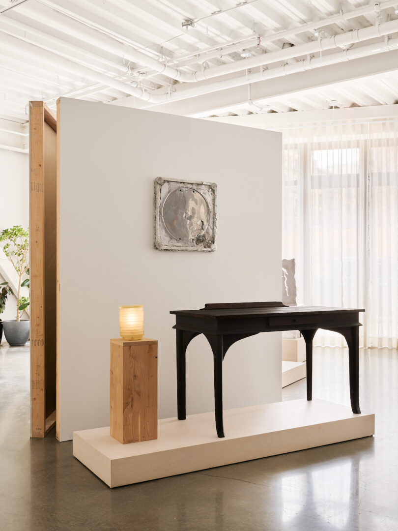A minimalist gallery space featuring a dark wooden desk on a platform, a wooden pedestal with a textured lamp, and an abstract artwork on a white wall