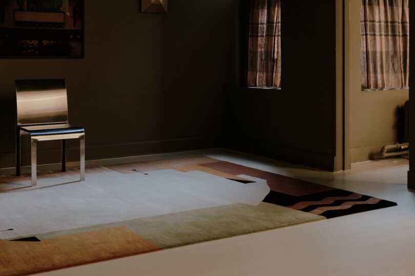 A metallic chair sits on a geometric-patterned rug in a dimly lit room with two windows covered by brown curtains.