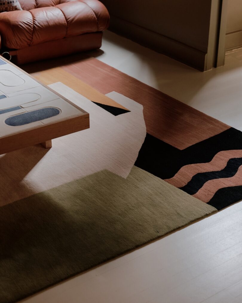 A modern living room with a geometric area rug, a wooden coffee table with a glass top, and part of a brown leather chair visible in the corner.