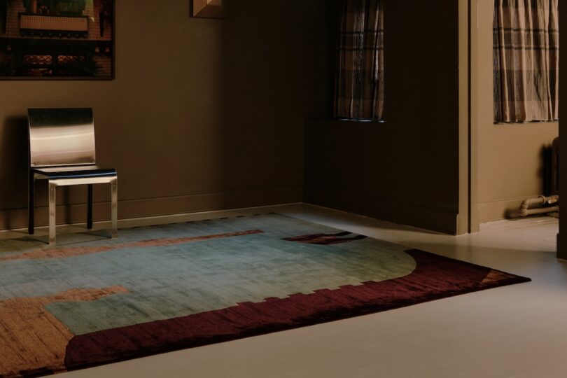A metallic chair sits against a brown wall in a sparsely furnished room with a patterned area rug and two curtained windows.