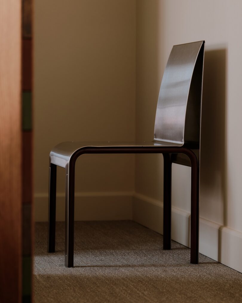 A modern, minimalist metal chair with a curved seat and backrest positioned against a beige wall on a carpeted floor.