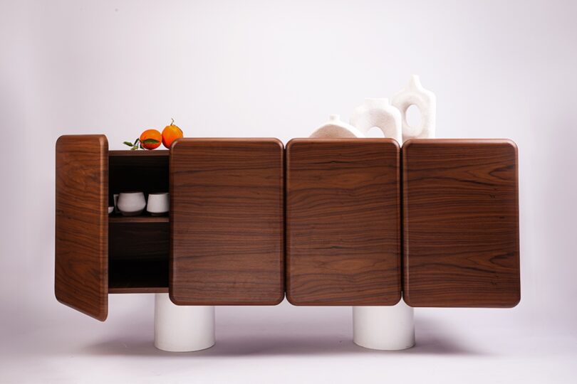 A wooden sideboard with four square doors, one slightly open revealing teacups inside. Three white vases and two oranges are placed on top