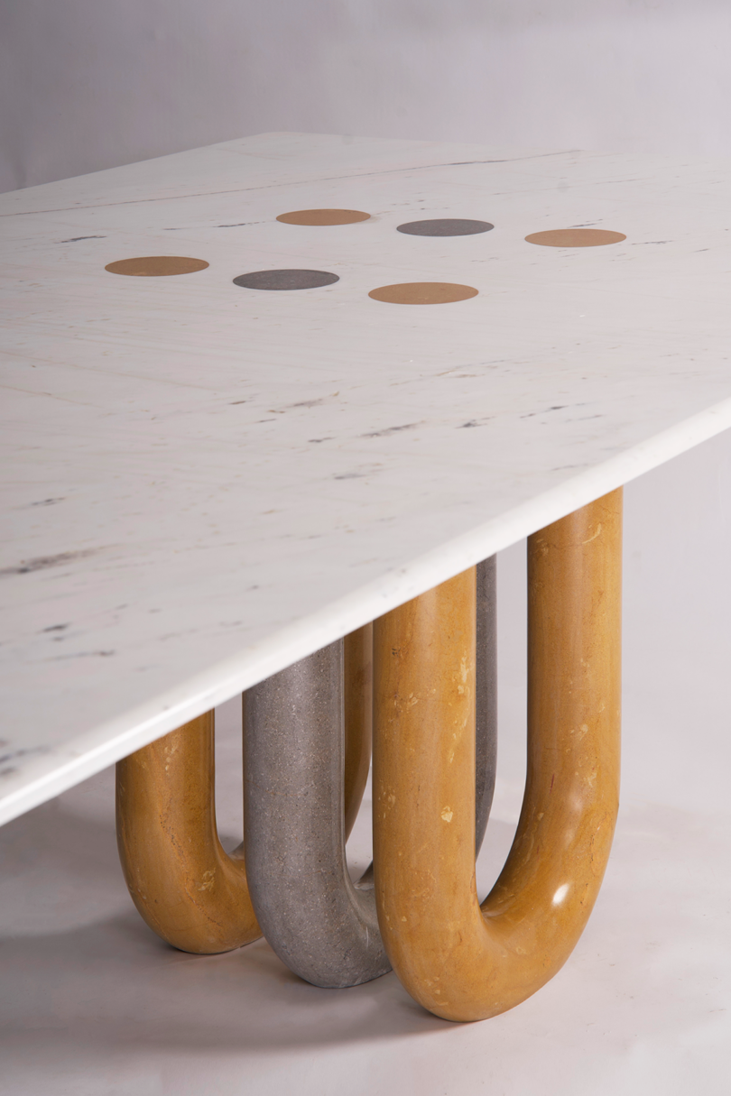 A white rectangular table with circular inlays on the surface. The table is supported by four large, curved, industrial-style legs in gray and brown