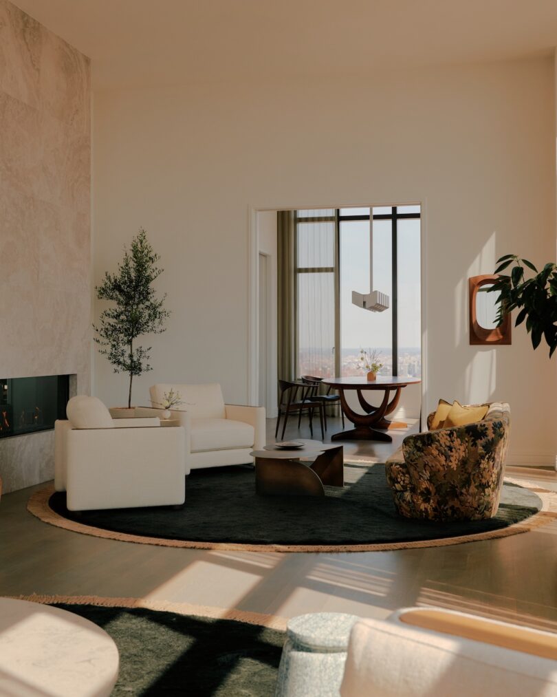 A modern living room with neutral-toned furniture, a round dark rug, large windows, indoor plants, and an adjacent dining area with city views.