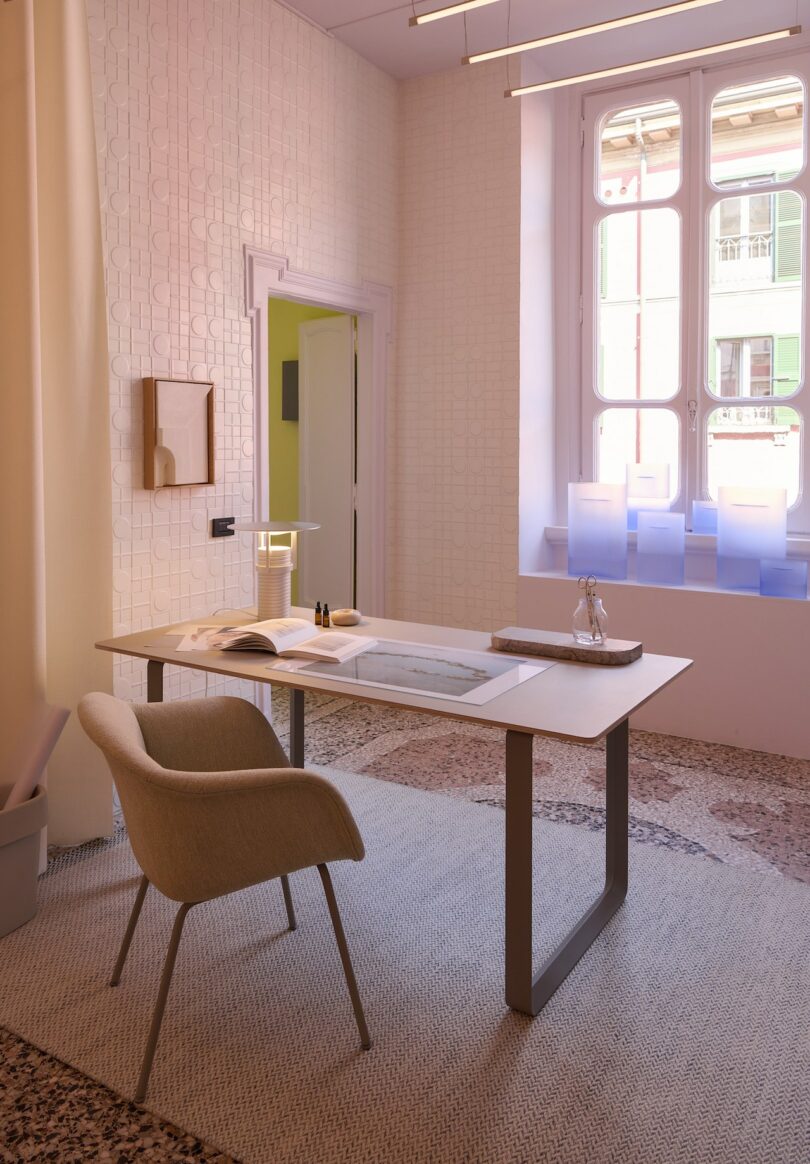 Modern minimalist office with a sleek desk, beige chair, open book, lamp, decorative items, tall window, and patterned floor. Light streams in through the window
