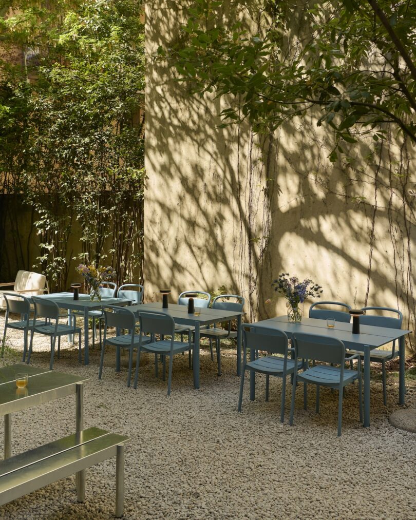 Outdoor patio with blue chairs and tables on gravel, shaded by trees and greenery, with vases of flowers and drinks on the tables