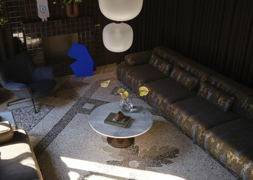 A modern living room with a large brown sectional sofa, a round coffee table holding a vase and book, an armchair, geometric wall art, and a tiled fireplace
