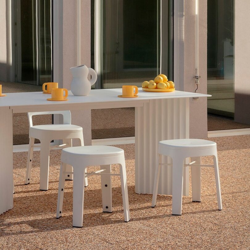 Outdoor patio with a white table and stools. A ceramic pitcher and four mugs are on the table. A bowl filled with lemons is at one end. Large windows reflect the surroundings