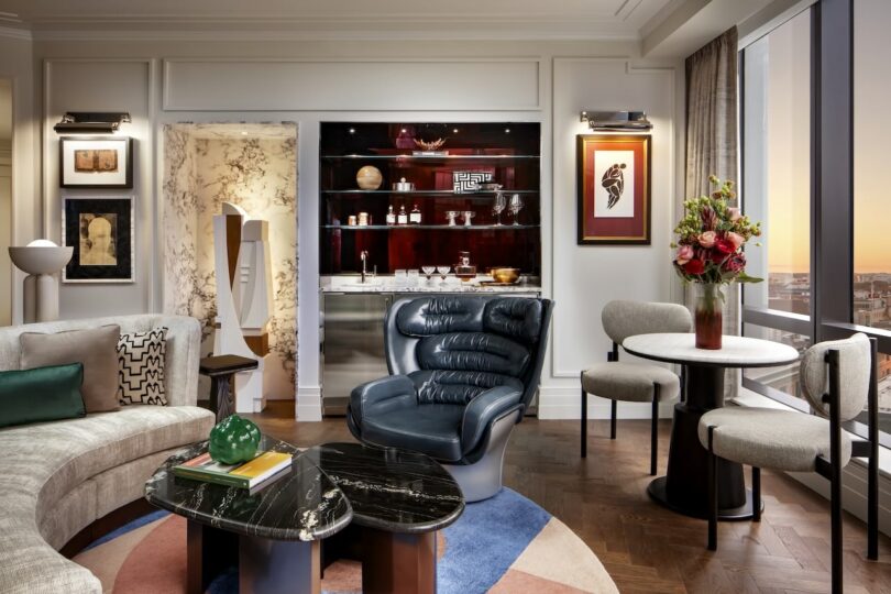 Modern living room with a curved sofa, designer armchair, marble coffee tables, and a small dining area. Bar area with shelves and art on the walls. Large windows offer city sunset views.