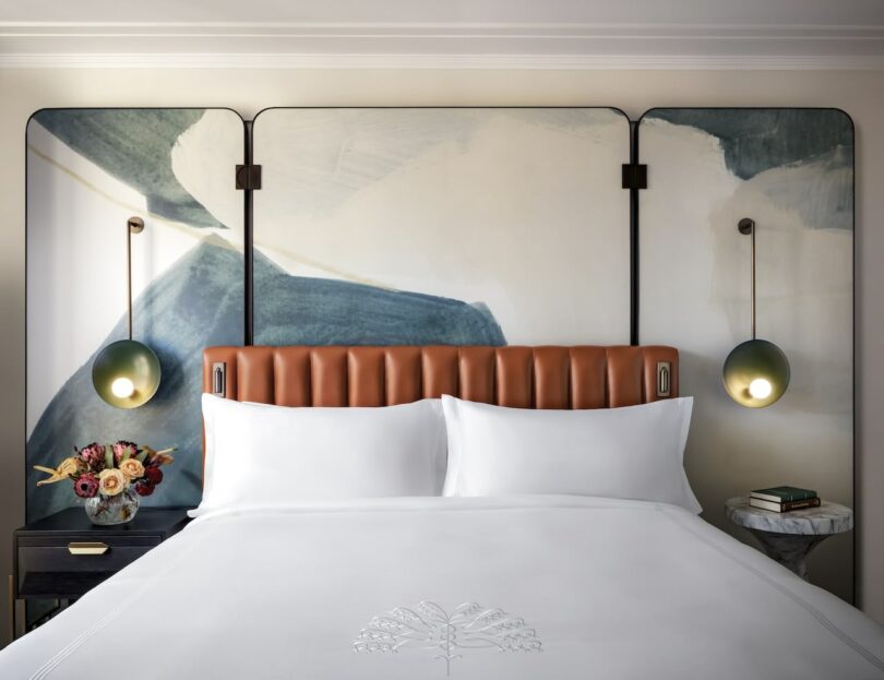 Modern bedroom with a leather upholstered headboard, abstract wall art, pendant lights, white bedding, a nightstand with flowers, and a small round table with books.