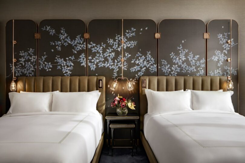 Two neatly made double beds with white bedding, separated by a nightstand with a vase of flowers. The headboard features a decorative panel with a floral design. Pendant lights hang above.