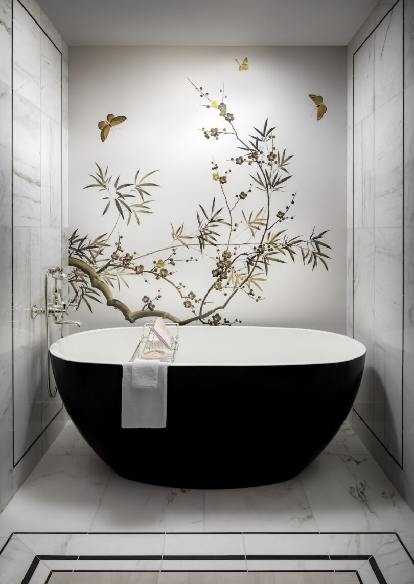 Elegant bathroom with a black freestanding bathtub, marble floors, and a wall featuring a nature-themed mural with branches and butterflies. A towel and book rest on the tub's edge.