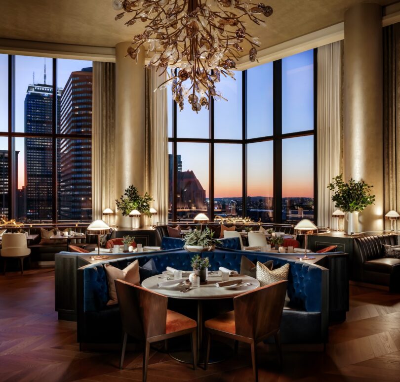 Elegant restaurant interior with large windows overlooking a cityscape at sunset. Tables are set with minimalistic decor, dark blue and brown seating, and a modern chandelier overhead.