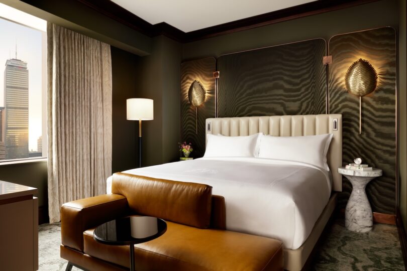 Modern hotel room with a large bed, leather sofa, and a view of a city skyline through the window. The room has dark green walls, a tall lamp, wall art, and a small marble-top side table.