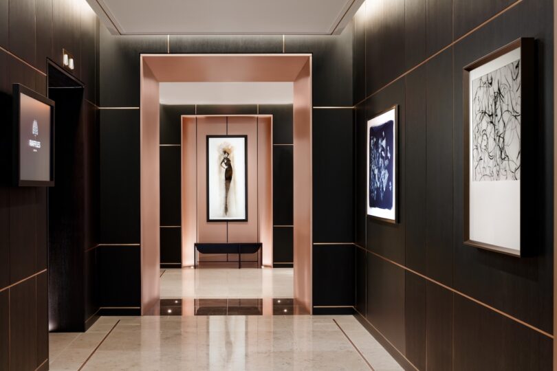 Hallway with dark paneled walls and framed art. Central lighted niche with a bench below an abstract painting. Marble floor and soft lighting create a minimalist, modern ambiance.