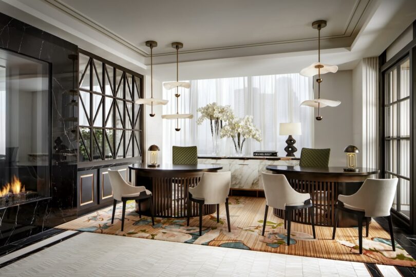 Luxurious dining room with two tables, six chairs, modern pendant lights, a marble sideboard with orchids, floor-to-ceiling windows, and a fireplace. Neutral tones and geometric patterns.