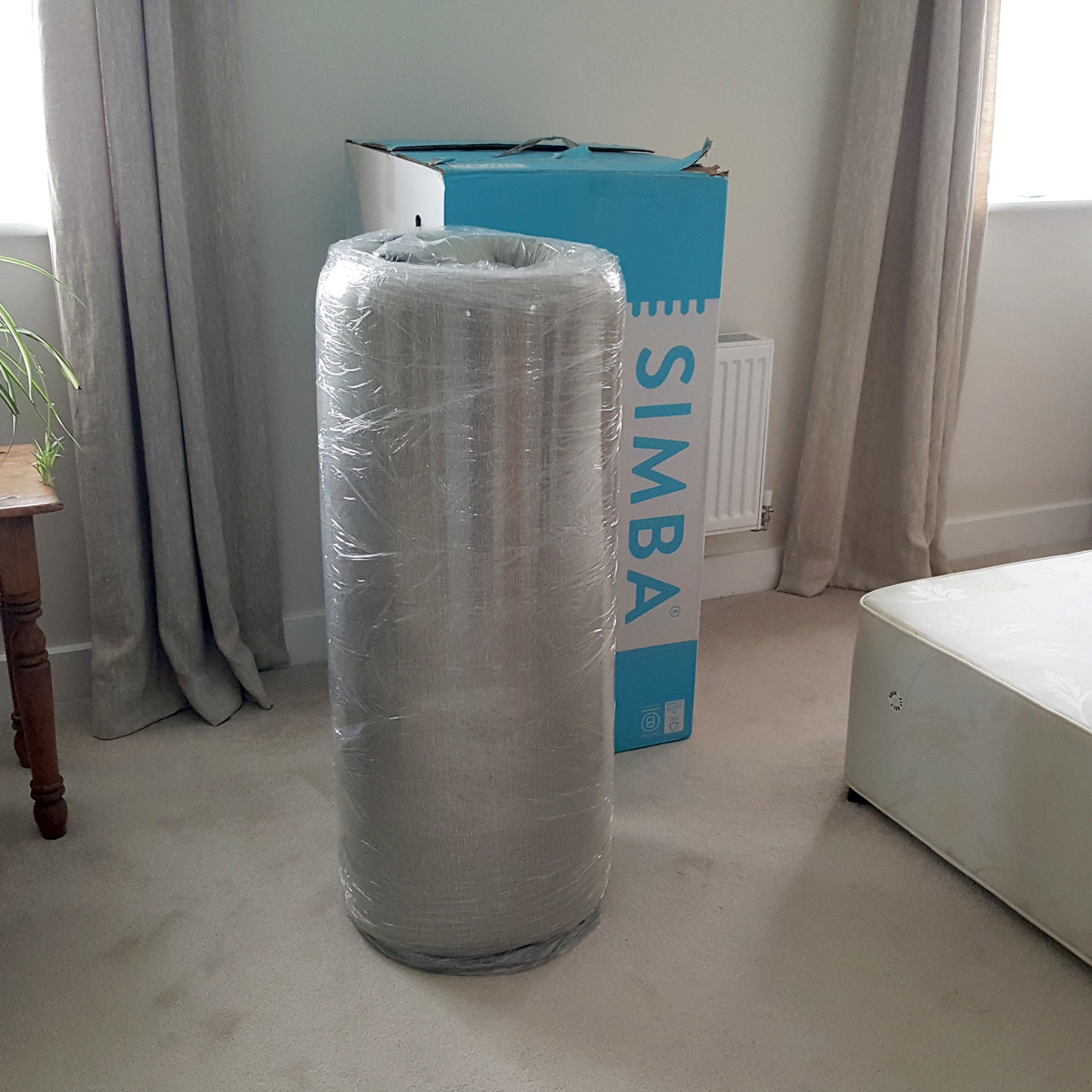 The Simba Hybrid Original mattress rolled up in a bedroom with its packaging box behind
