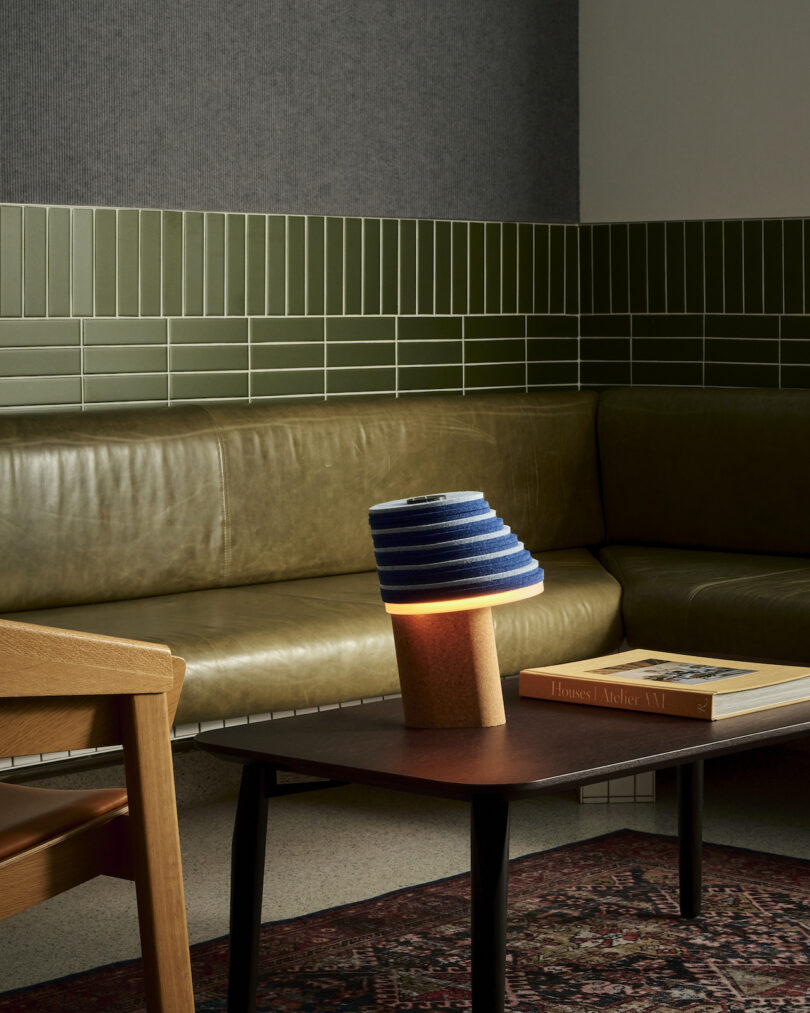 A modern living room with a green leather couch, a stackable green tiled wall, a striped lamp on a wooden coffee table, and a book placed next to the lamp