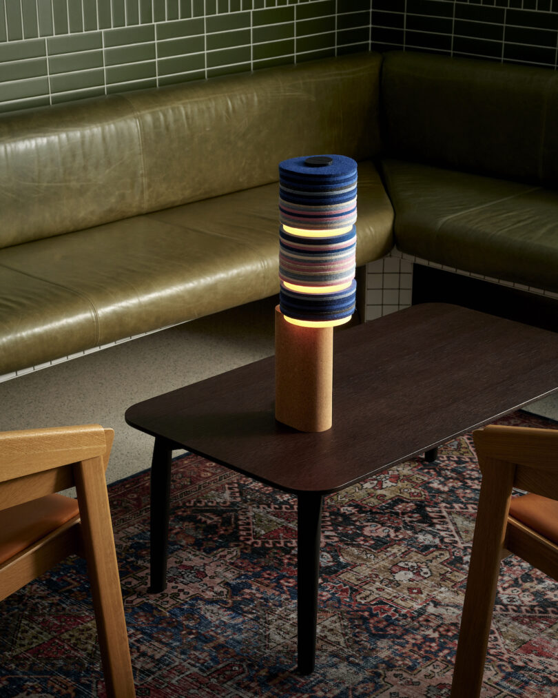 A modern cylindrical lamp with multicolored layers from the Stackabl collection sits on a dark wood table. The table is surrounded by green leather seating and wooden chairs atop a patterned rug