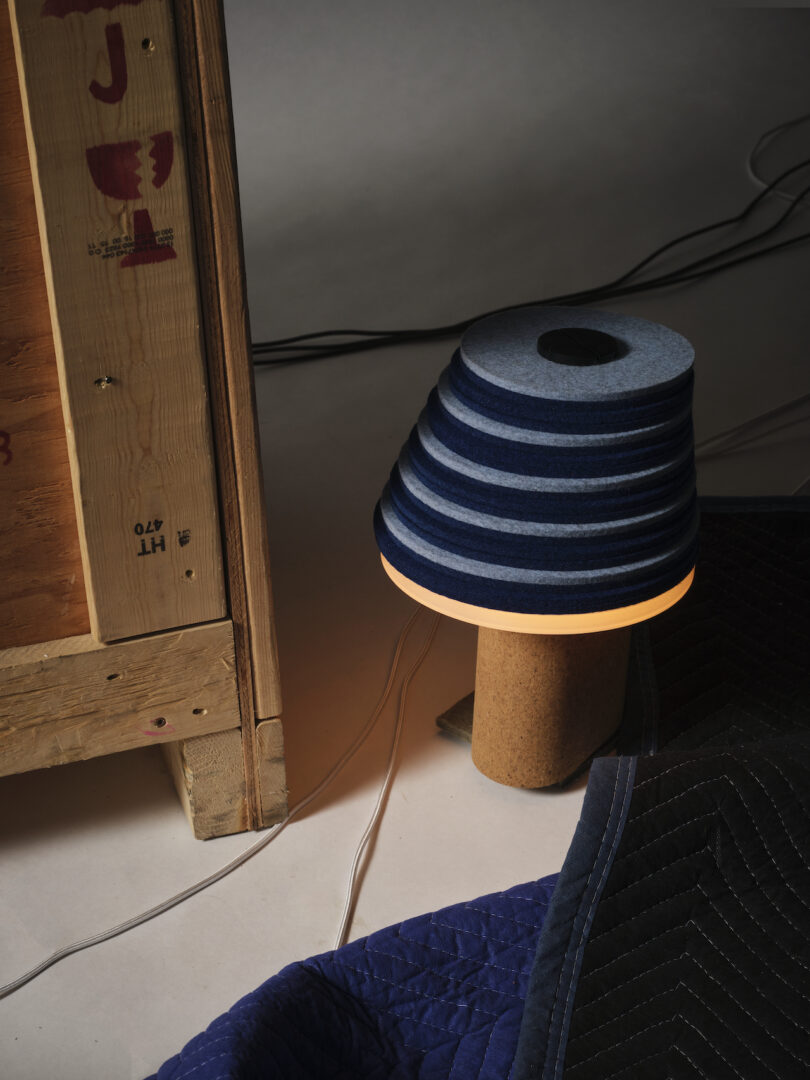 A striped blue and gray lamp with a wooden base, crafted using Stackabl techniques, is illuminated next to a wooden crate. Wires and a dark quilted fabric are on the floor