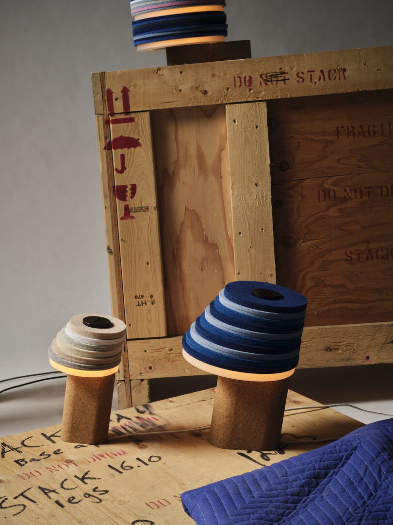 Three Stackabl lamps with layered, colorful shades and cork bases are artfully arranged on a wooden crate and platform. Blue padded fabric enhances the foreground