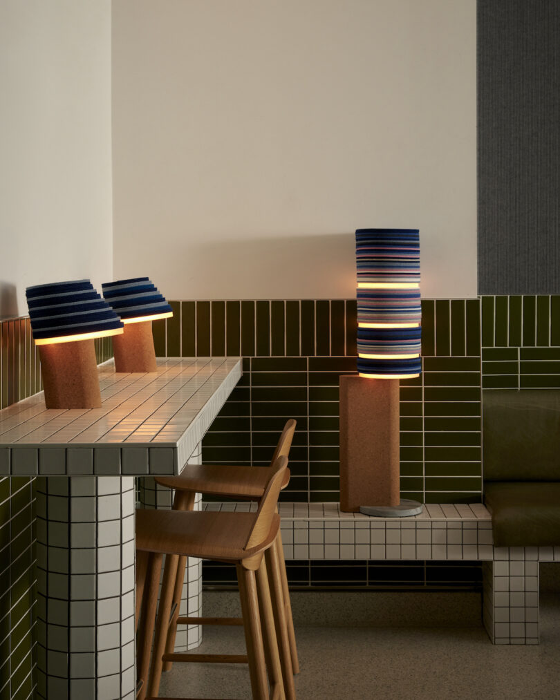 A cozy corner seating area features a Stackabl tiled table, two wooden chairs, and a cushioned bench. Three modern lamps with stripy shades cast a warm glow, enhancing the charm of the green-tiled walls