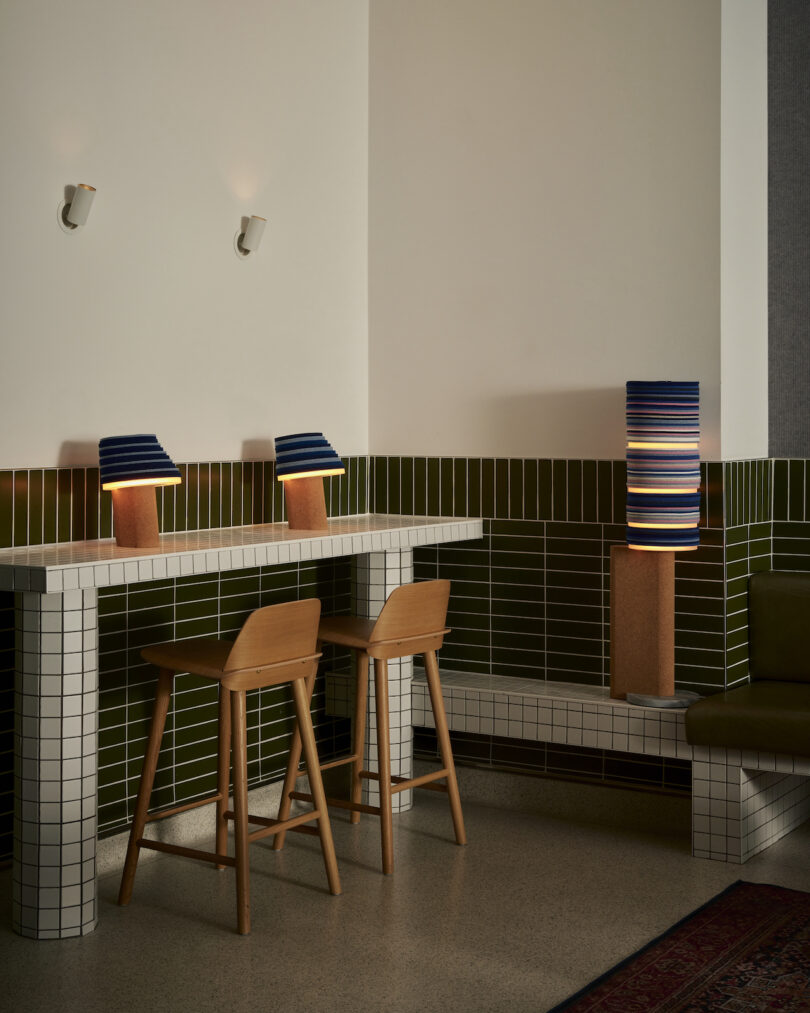 A modern interior featuring a Stackabl-inspired tiled bar, complemented by two wooden stools and contemporary lamps on both the counter and floor; green and white tiles artfully line the walls