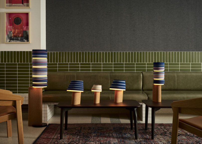 Cozy seating area with green tile and gray walls. Felt and cork lamps with blue and striped shades. Wooden chairs and a patterned rug complete the decor.