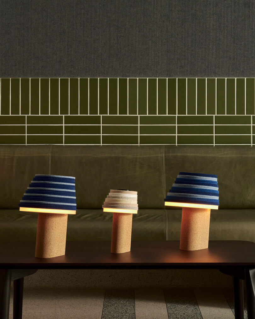 Three Stackabl table lamps with cork bases and tiered, colorful shades sit on a wooden table against a green tiled wall and leather bench