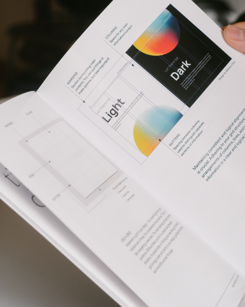 A person holds open a book showing a design guide with diagrams comparing light and dark layouts, grid lines, and labeled sections