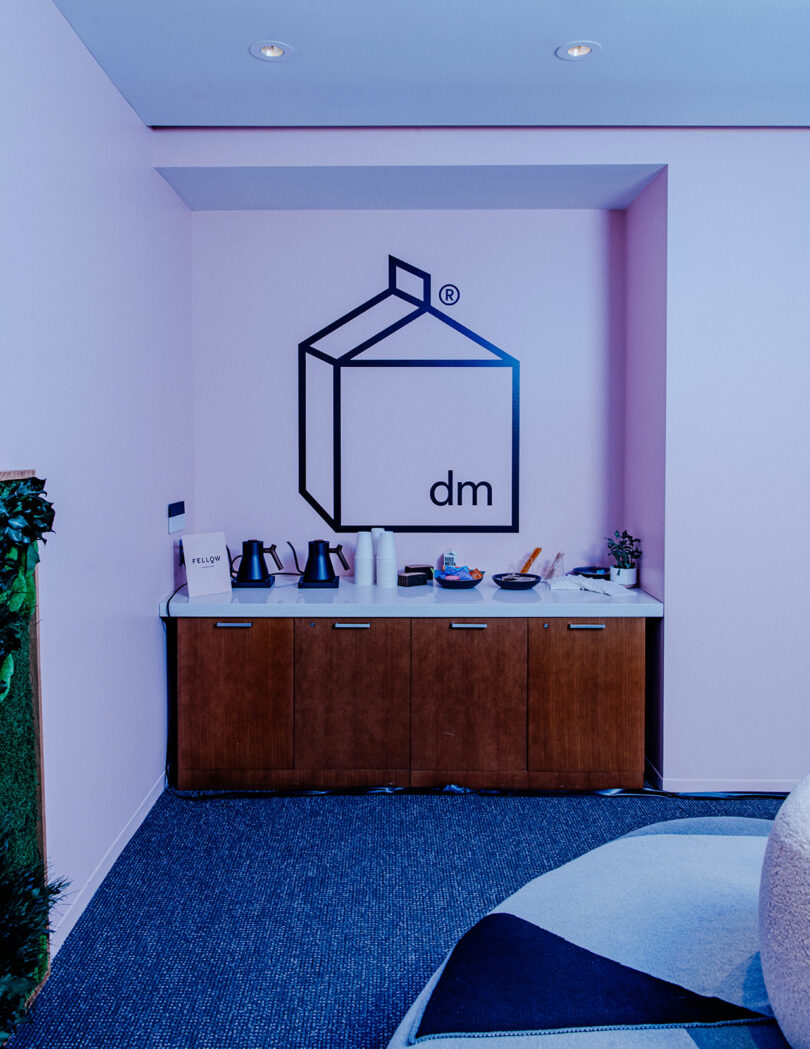 A small refreshment station with kettles, cups, and snacks sits against a pink wall with a geometric logo and the letters "dm" above it, evoking the cozy ambiance of a Designer Wellness Lounge.