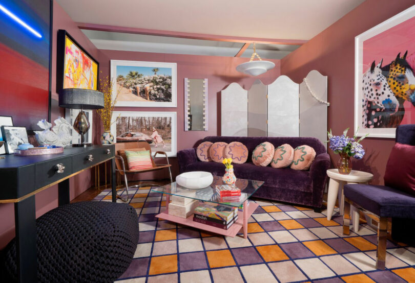 A colorful living room by Tina Ramchandani features a purple sofa, patterned rug, pink walls, art on the walls, a black ottoman, a console table, and a coffee table with books and decorative objects.