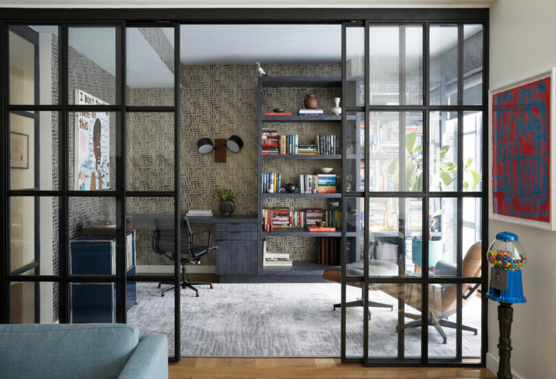 Modern home office with glass sliding doors, a desk, shelves with books, and large windows letting in natural light. Designed by Tina Ramchandani, the space features art, an armchair, and a gumball machine in the foreground.